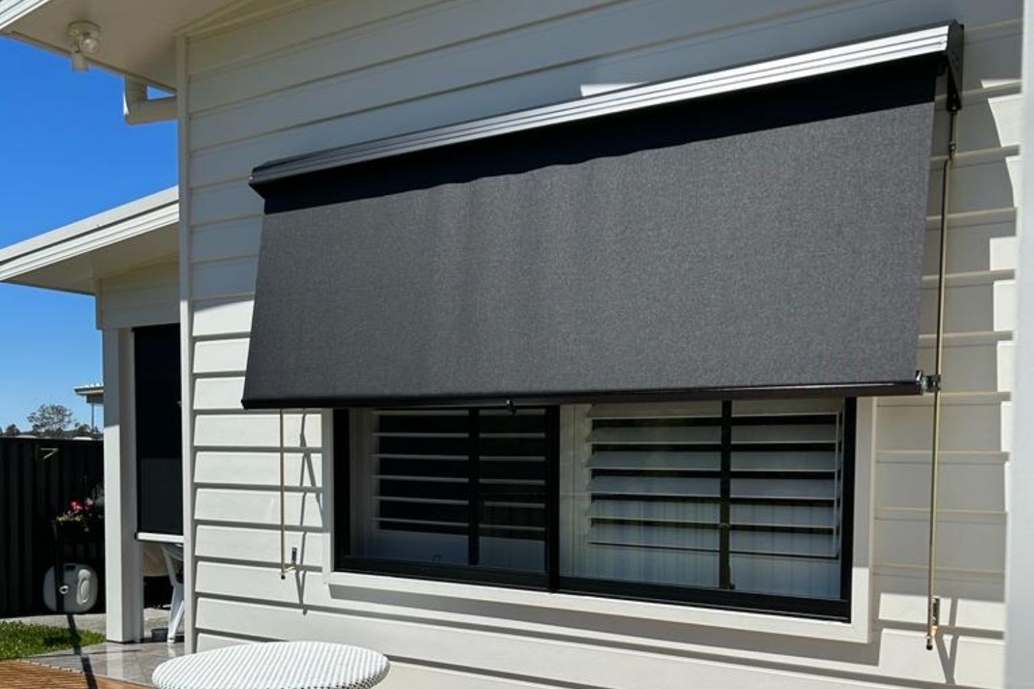 Black residential window awning installed above a black-framed window on a white-clad Wollongong home with a timber-look deck.