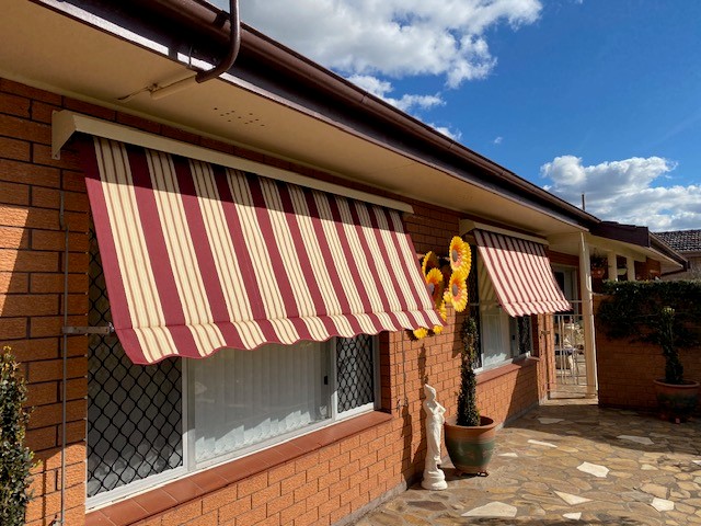 Striped automatic awnings installed over front windows of a brick home in Greater New England by Apollo Blinds